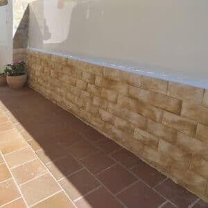 Un revestimiento de pared exterior con ladrillos de piedra marrón claro en una base blanca y suelo de baldosas rojas, con macetas de flores en el lateral.