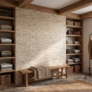 Espacio interior con revestimiento de piedra beige en una pared, suelo de madera y estanterías de madera con ropa y accesorios, banco de madera con manta.