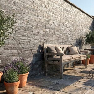 Espacio exterior con pared de piedra gris estilo rústico, banco de madera y plantas en macetas de terracota, suelo de piedra.