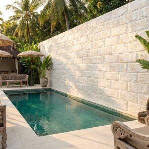 Espacio exterior con piscina rectangular de agua turquesa rodeada de suelo y pared de piedra clara, sillas de madera tallada y palmeras al fondo.