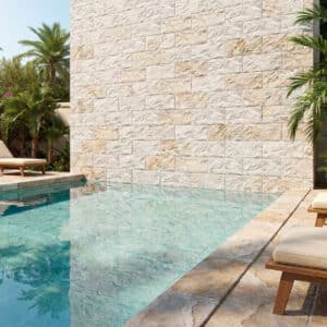 Un espacio de piscina al aire libre con suelo y pared de piedra clara, adornado con sillas de madera y plantas tropicales.